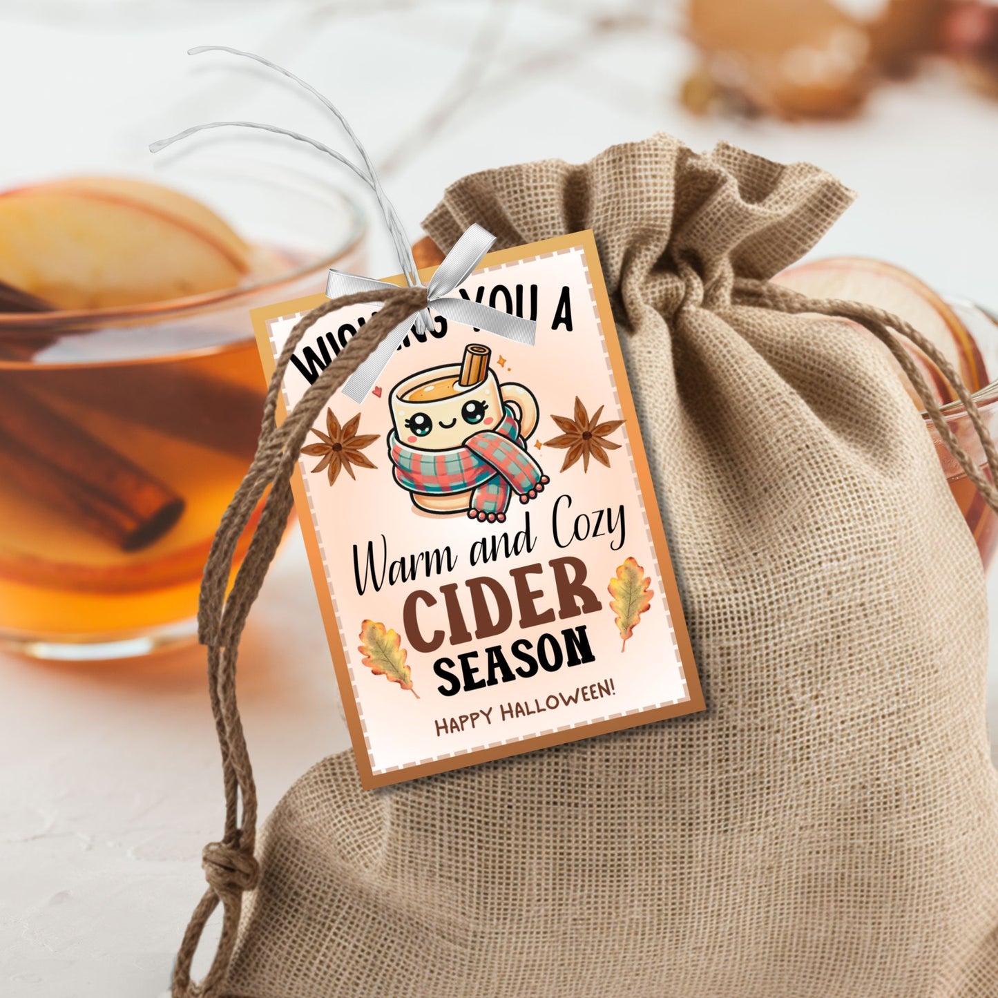 A printable Halloween gift tag featuring a warm and cozy cider theme. The tag displays a cute mug wrapped in a scarf, surrounded by autumn leaves and star anise, with the message "Wishing You a Warm and Cozy Cider Season."