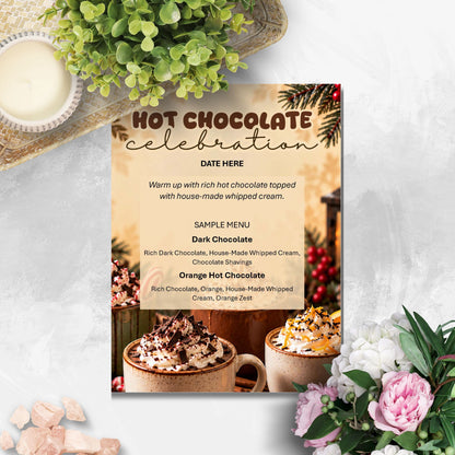 A printed hot chocolate celebration menu card styled on a light marble surface with flowers, a candle, and greenery around it. The card features a warm beige center, dark brown header text reading 'HOT CHOCOLATE celebration,' sample menu text, and photos of chocolate-dusted hot chocolate mugs at the bottom.