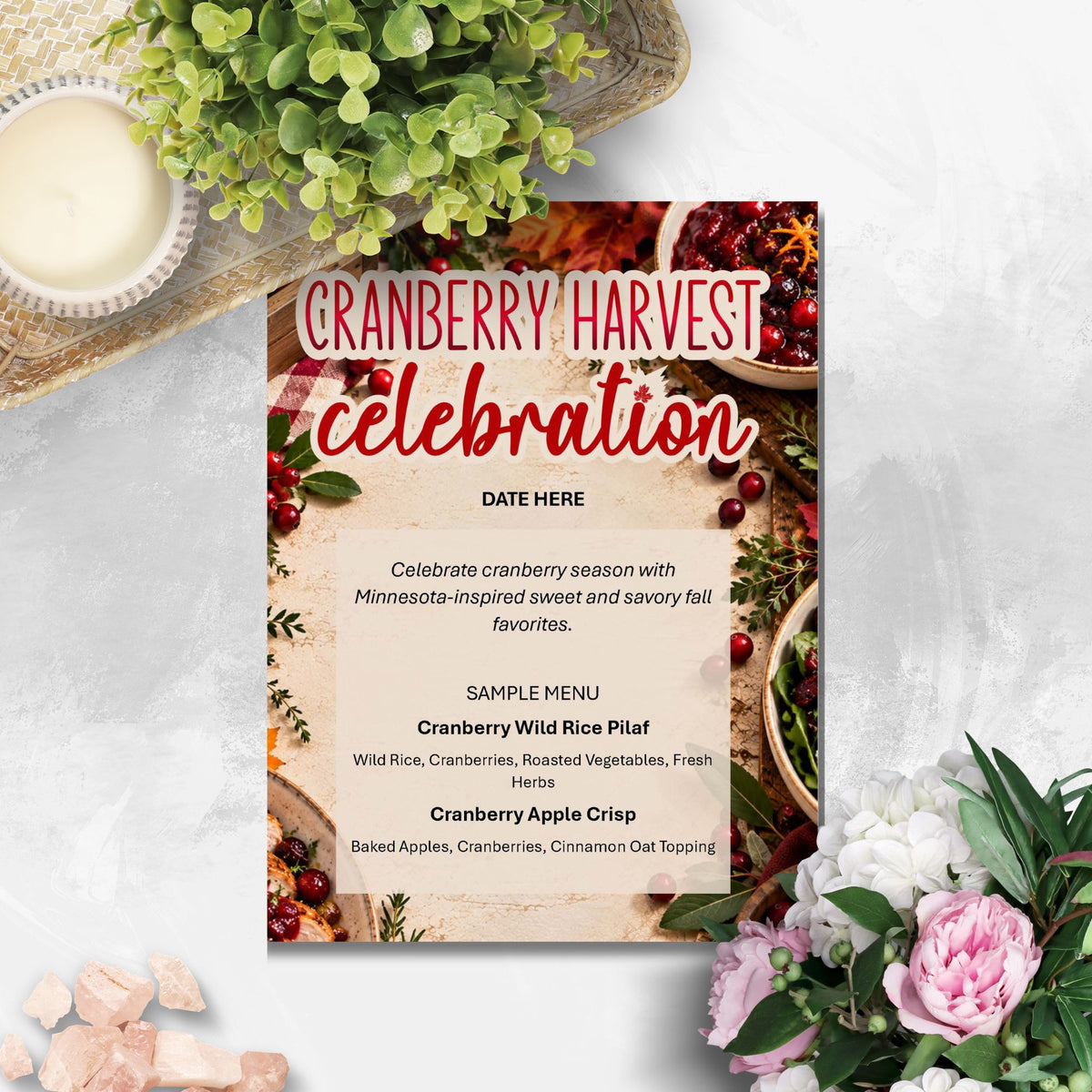 A printed invitation card styled on a light marble surface with decorative greenery, flowers, and small stones around it. The card reads 'CRANBERRY HARVEST celebration' and shows a sample menu for fall foods.
