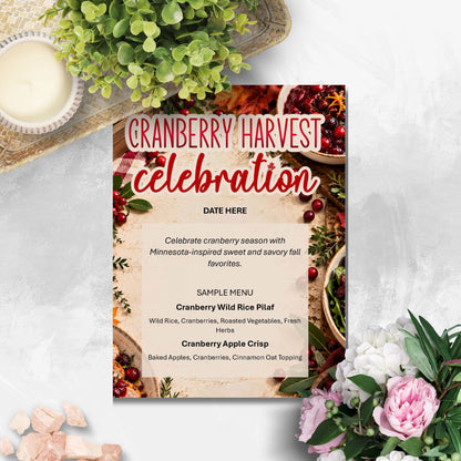 A printed invitation card styled on a light marble surface with decorative greenery, flowers, and small stones around it. The card reads 'CRANBERRY HARVEST celebration' and shows a sample menu for fall foods.