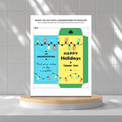 A printed holiday housekeeping tip envelope template displayed upright on a round pedestal against a tiled wall. The card design shows a split teal and yellow holiday gift box with colorful string lights and text reading 'Happy Holidays & Thank You!' and 'Thank you for making our stay so comfortable!'.