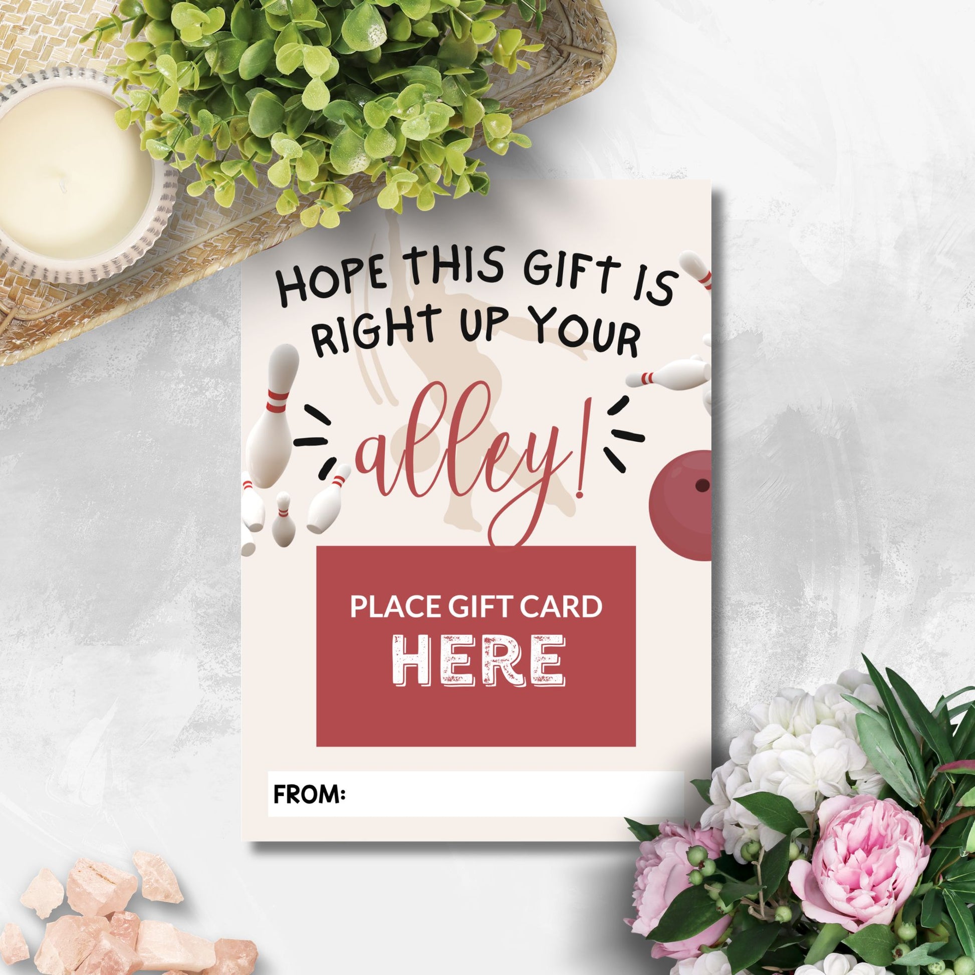 A novelty bowling-themed gift card with the text 'HOPE THIS GIFT IS RIGHT UP YOUR alley!' and a red box reading 'PLACE GIFT CARD HERE', styled on a light marble surface with flowers, a candle, and greenery around it.