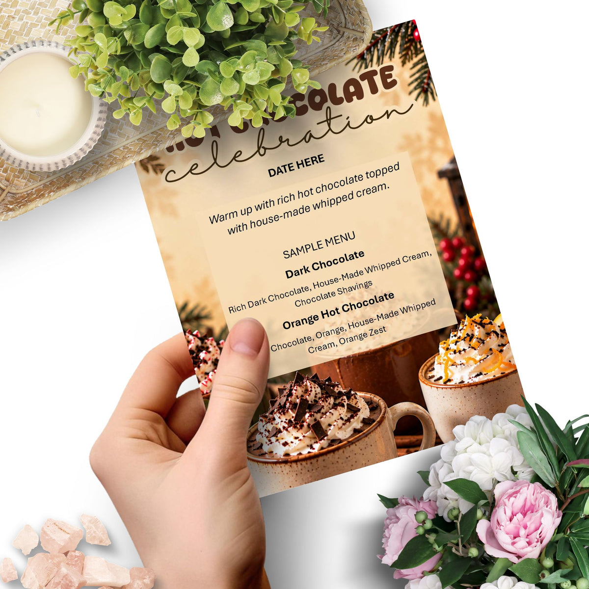 A hand holds a printed hot chocolate celebration menu card over a styled tabletop scene with flowers, a candle, and mugs of chocolate drink.