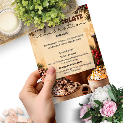 A hand holds a printed hot chocolate celebration menu card over a styled tabletop scene with flowers, a candle, and mugs of chocolate drink.