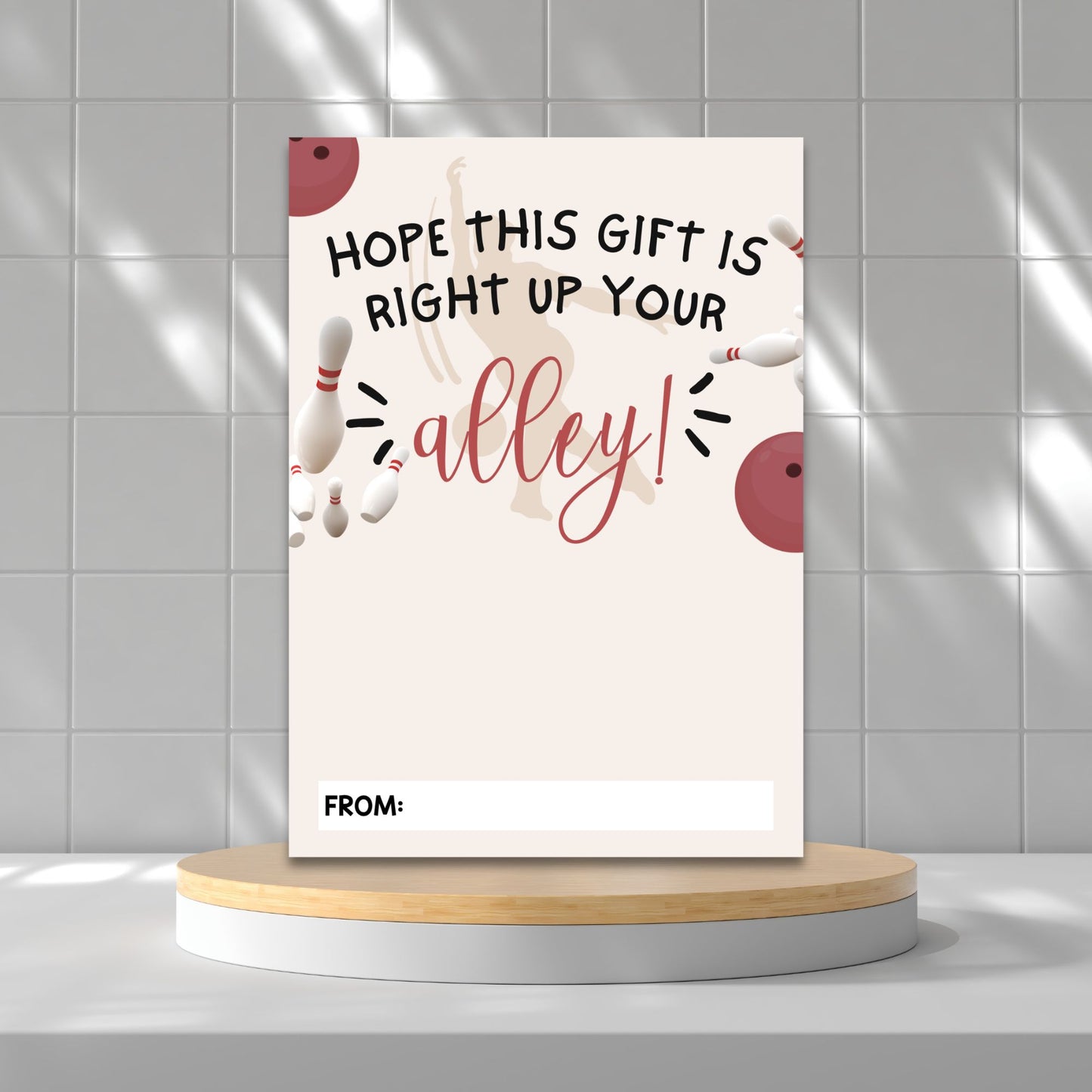 A rectangular gift card with bowling-themed graphics and the text 'HOPE THIS GIFT IS RIGHT UP YOUR alley!' stands upright on a round wooden platform against a light gray tiled wall.