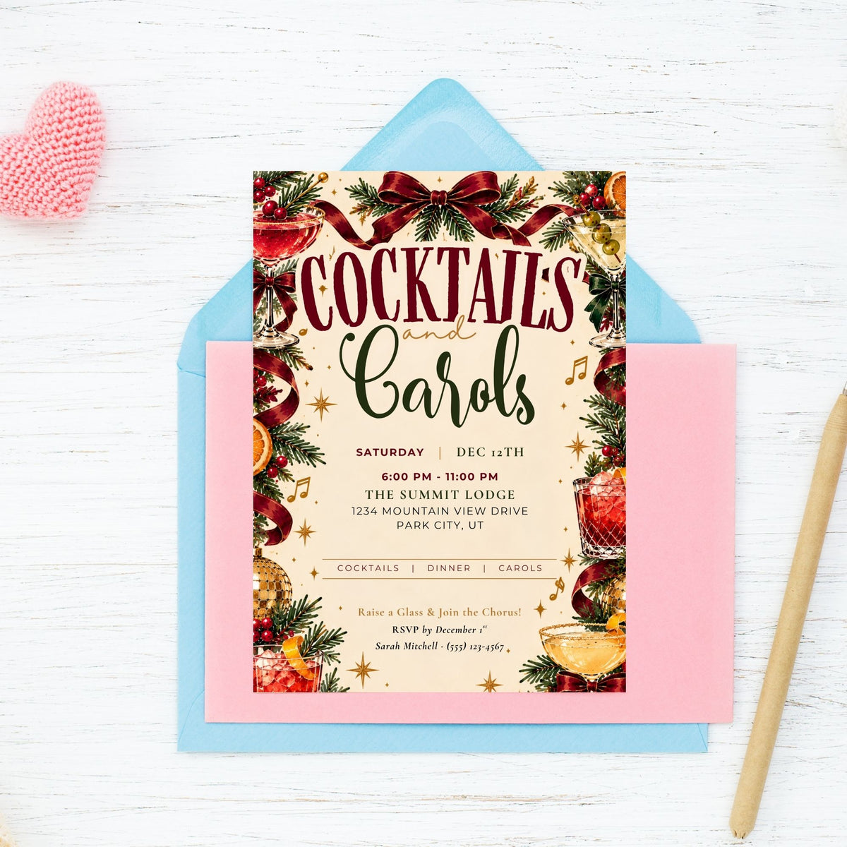 A festive 'Cocktails and Carols' cocktail party invitation card displayed on top of a light blue envelope and a pink card, arranged on a white wood background with a pink heart-shaped ornament and a gold pen.