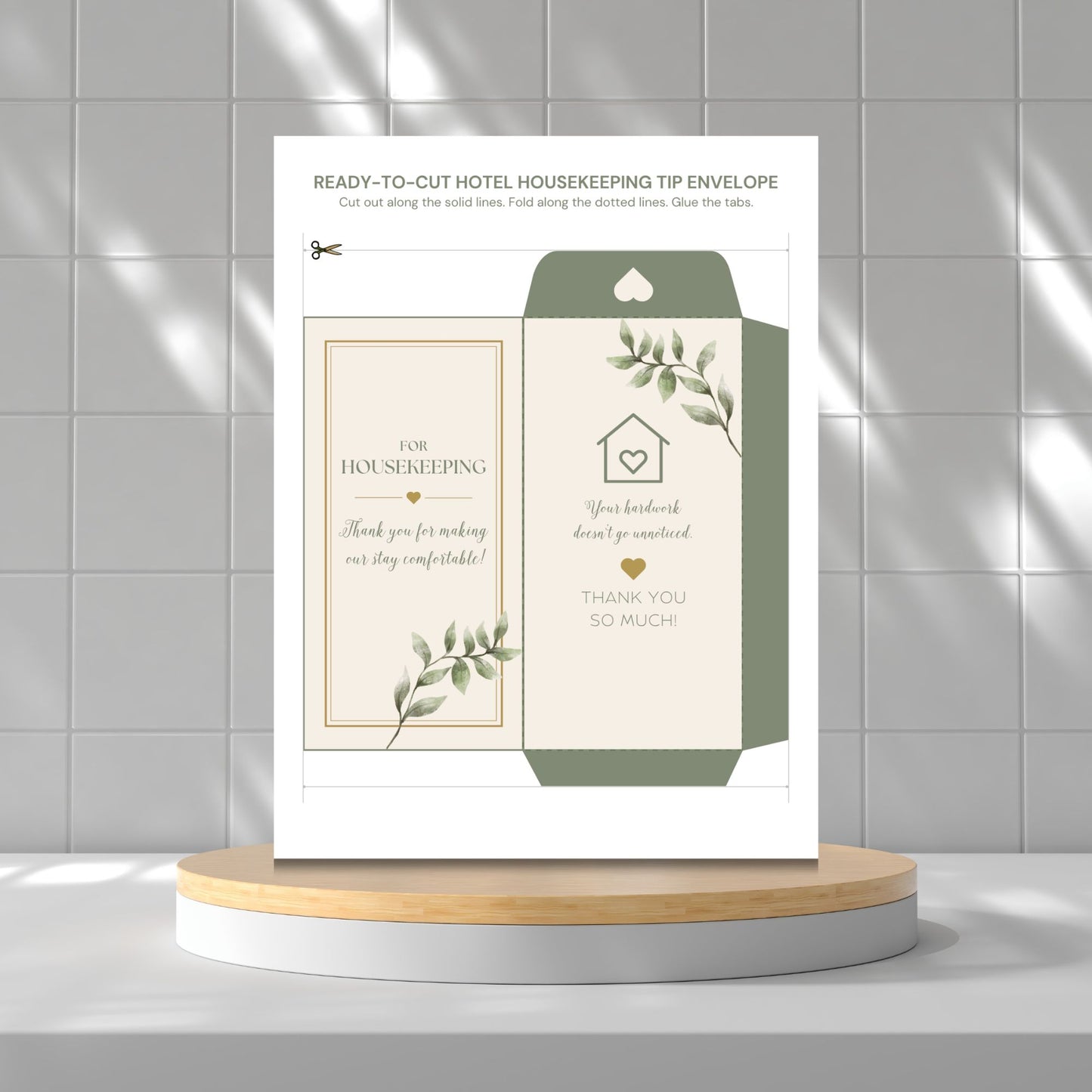 A printed housekeeping tip envelope template shown upright on a round pedestal against a tiled wall. The envelope design is cream and sage green with botanical leaf illustrations and placeholder text for housekeeping thanks and compliments.