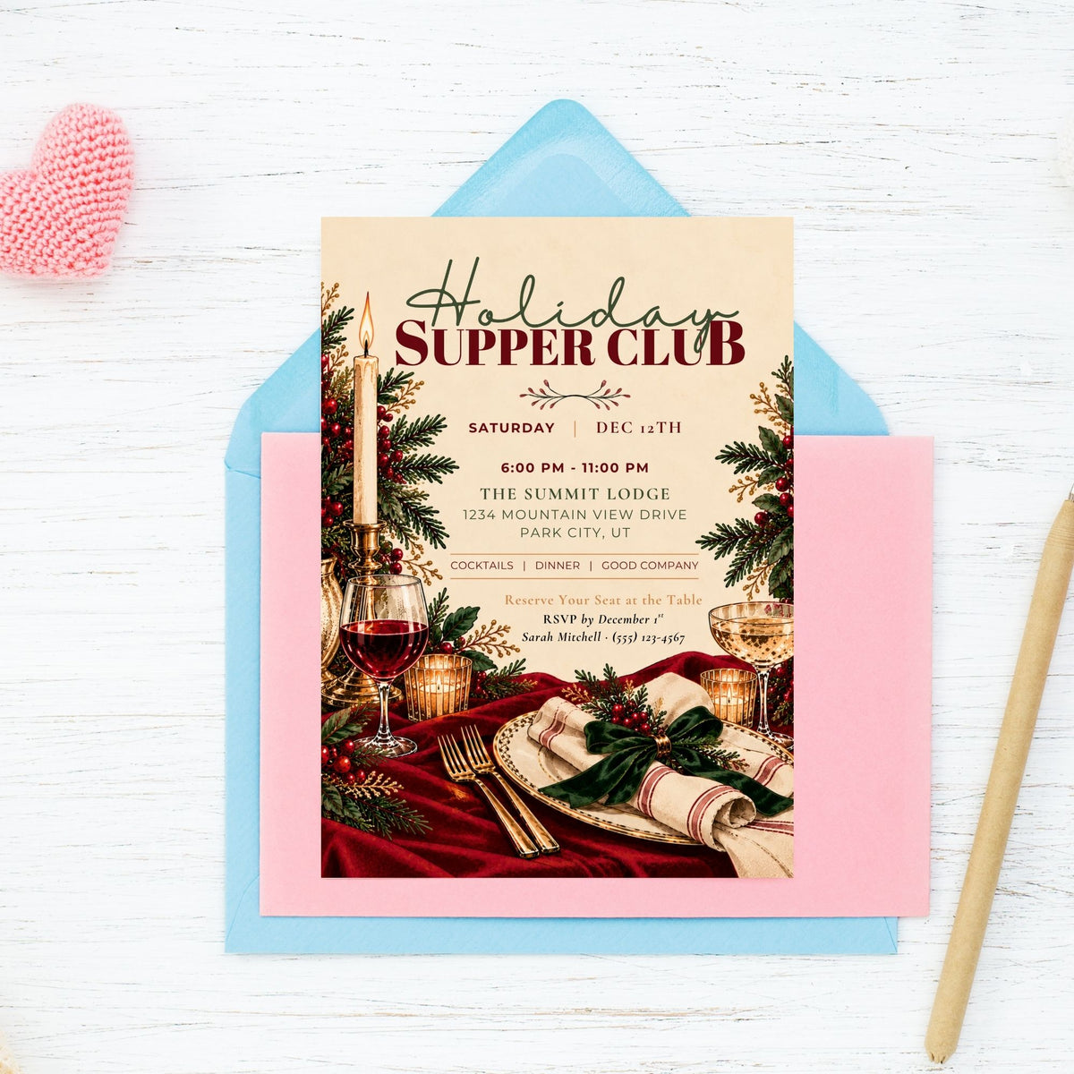 A styled flat-lay of a 'Holiday Supper Club' invitation card featuring a festive dinner scene, layered over a light blue envelope and a pink card, set on a white wood background with a pink heart and gold pen.