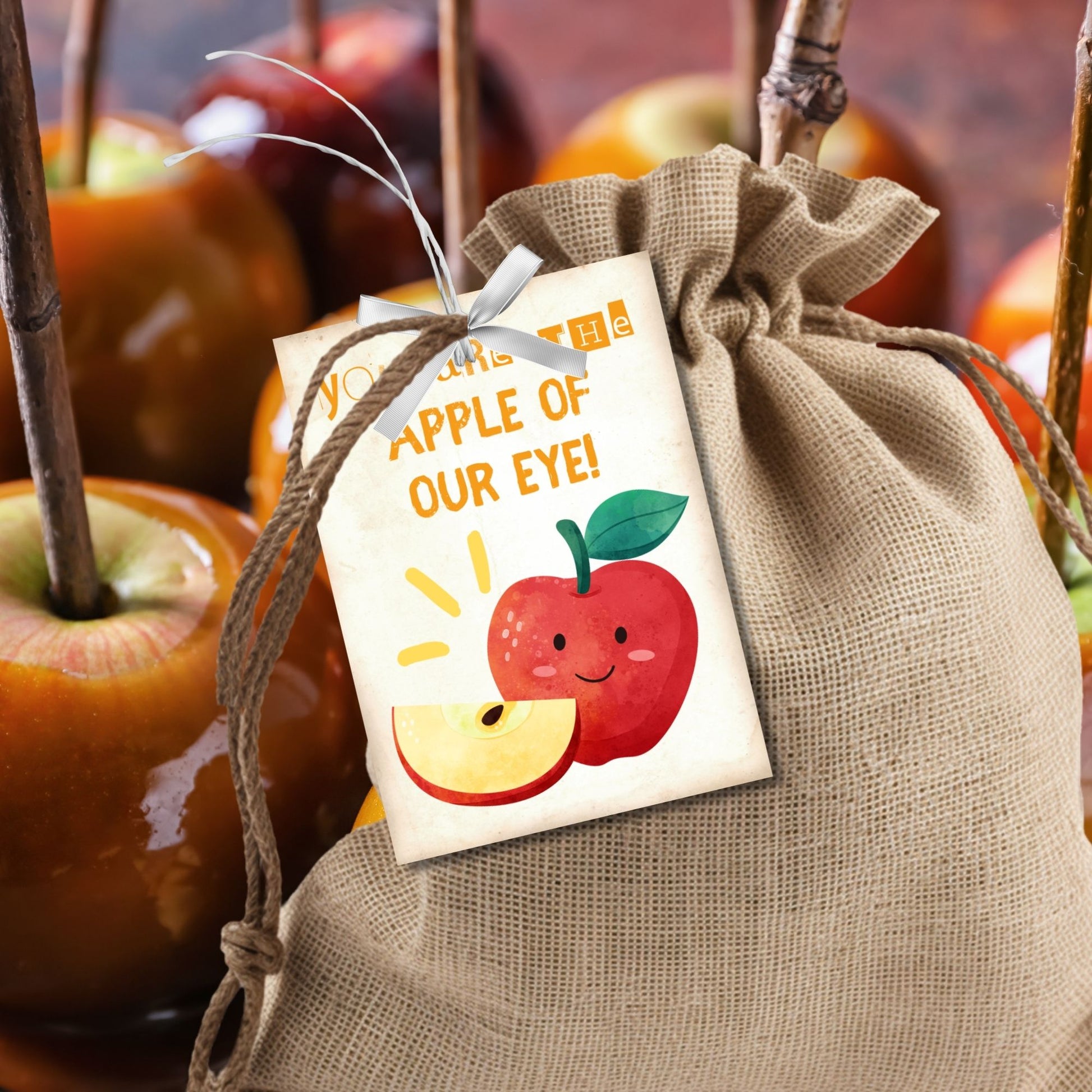 Halloween-themed printable gift tags with the message "You Are the Apple of Our Eye!" featuring a cute apple character perfect for caramel apple gifts. Tags are 2.5 x 3.5 inches, laid out 8 per sheet on a standard 8.5 x 11-inch page. Includes a printable PDF and a PDF with a link to an editable Canva template.