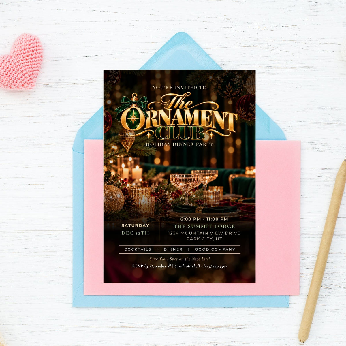 A holiday dinner party invitation card for 'The Ornament Club' is displayed on top of a light blue envelope and a pink card, set on a white wood background with a pink heart and a gold pen as props.