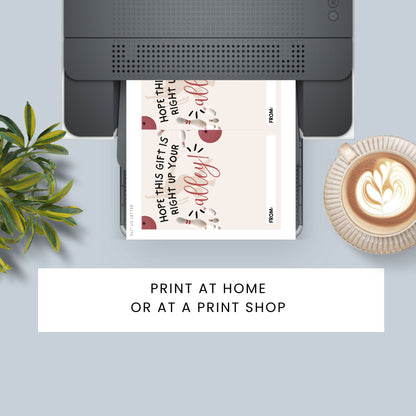 A top-down styled flat lay showing a printer with a printed gift tag sheet emerging, alongside a plant, a cup of latte, and a text banner reading 'PRINT AT HOME OR AT A PRINT SHOP'. The gift tag has a cream background with red and black decorative text and illustrations.