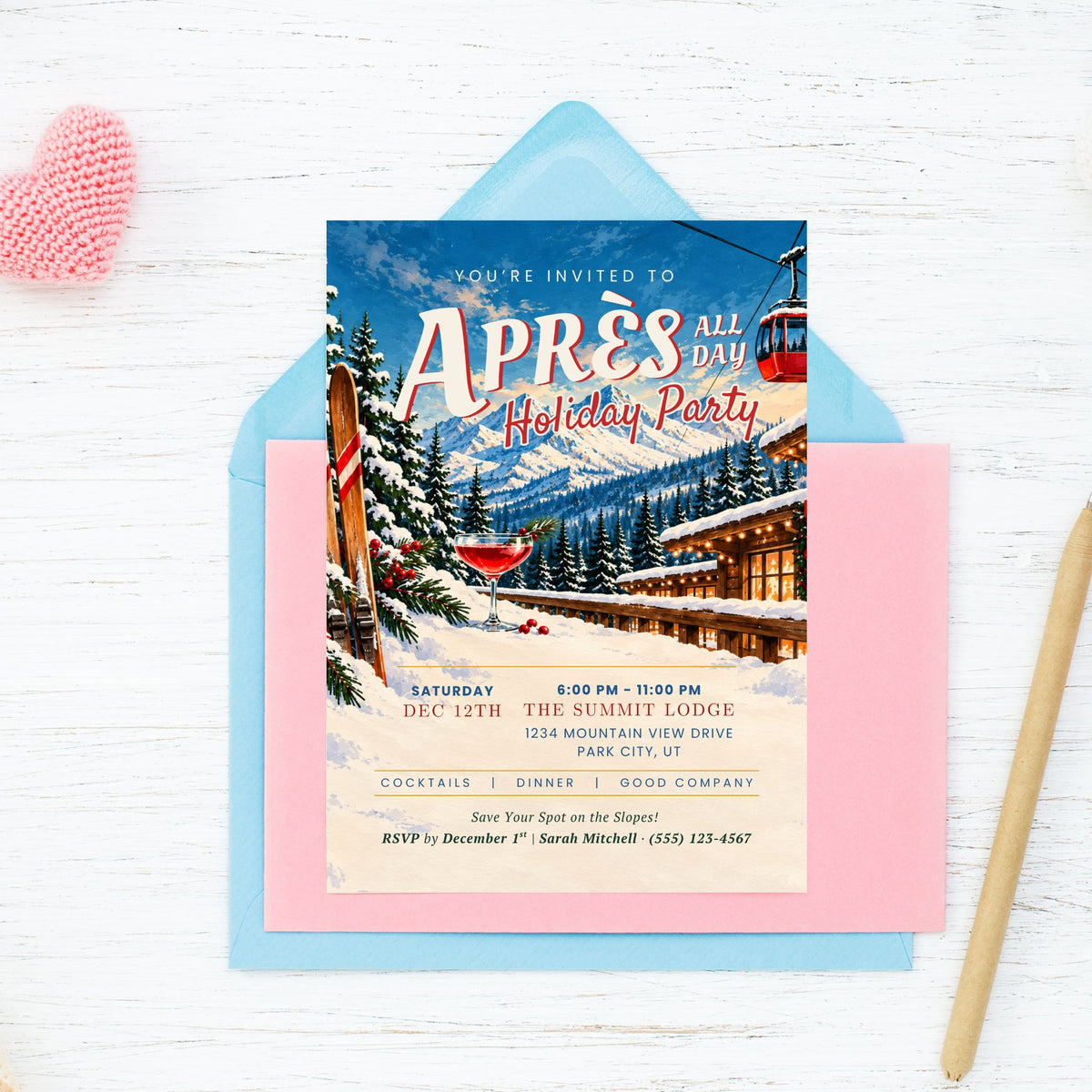 A winter-themed holiday party invitation card displayed on top of a light blue envelope and pink cardstock, styled on a white wood background with a pink heart and a wooden pencil.