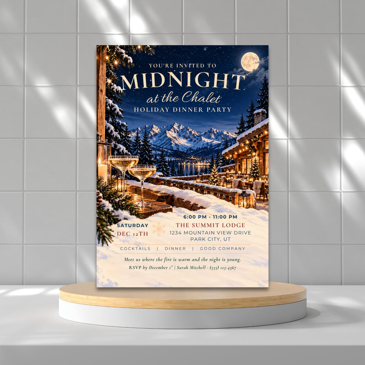 A vertical holiday dinner party invitation card displayed on a round wooden pedestal against a light gray tiled wall. The invitation features a winter mountain lodge scene at night with snow-covered trees, warm glowing windows, a full moon, and elegant white text reading 'Midnight at the Chateau Holiday Dinner Party.