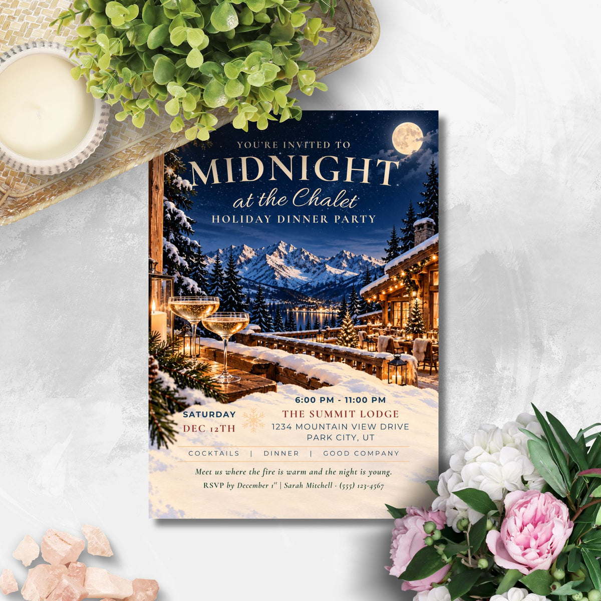A winter-themed dinner party invitation card styled in a flat-lay arrangement on a light gray marble surface, surrounded by a green plant, a lit candle, pink stones, and pink flowers. The card features an illustration of a snowy mountain lodge at night with a full moon and a hot air balloon.