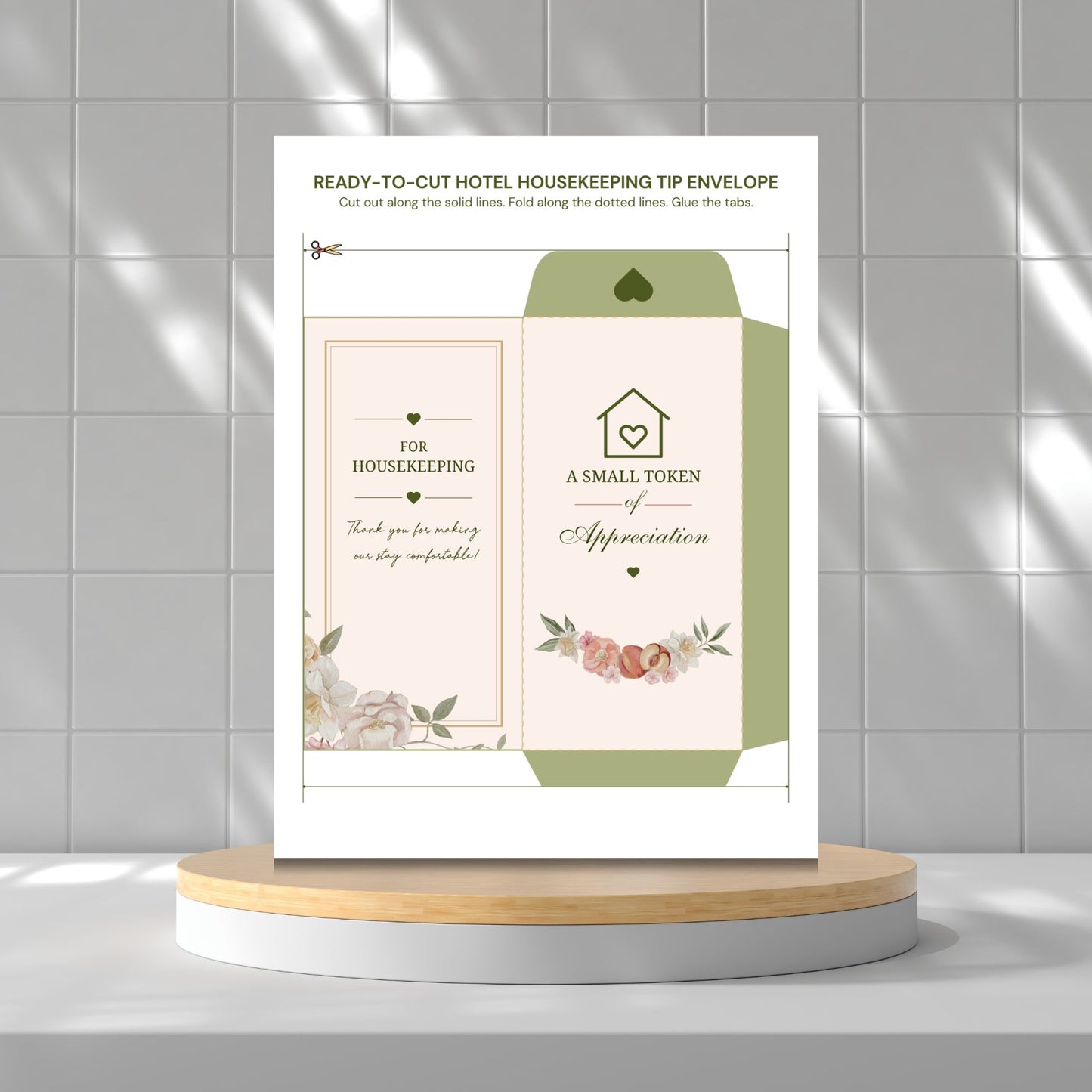 A printed housekeeping tip envelope template card displayed upright on a round wooden pedestal against a light gray tiled wall. The card shows a green envelope with two cream insert panels labeled for housekeeping and a small appreciation token, with floral illustrations at the bottom.