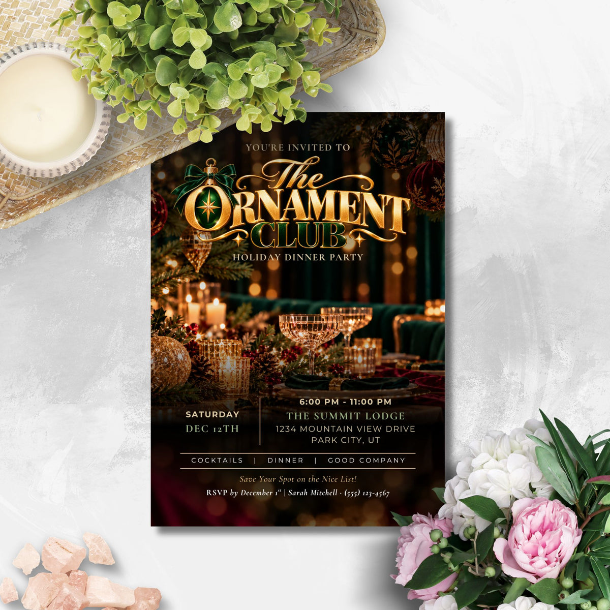A holiday-themed dinner party invitation card styled in a flat-lay arrangement with green foliage, a lit candle, and pink and white flowers on a light gray textured surface. The invitation features a festive table setting with gold lettering that reads 'The Ornament Club' and 'Holiday Dinner Party'.