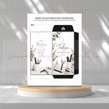 A printed hair stylist tip envelope card displayed upright on a round pedestal against a light gray tiled wall. The card shows a black-and-white envelope design with botanical leaf accents and small salon tools, plus text including 'READY-TO-CUT HAIR STYLIST TIP ENVELOPE' and 'A Small Token of APPRECIATION'.