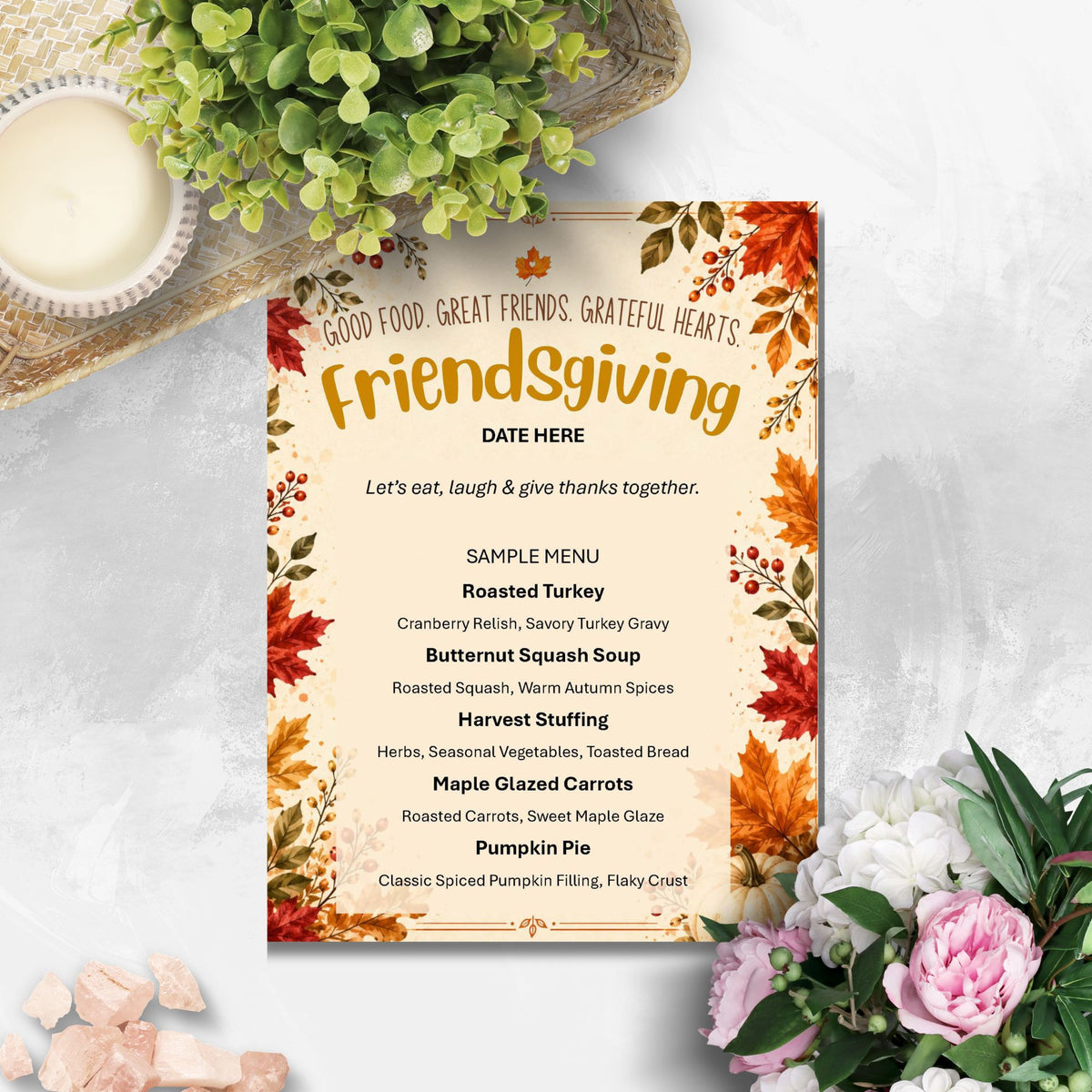 A Friendsgiving menu card styled in a flat-lay arrangement with autumn leaves, a green plant, a candle, and pink flowers on a light marble surface.