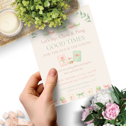 Hand holding a pink invitation card with floral decorations on a white surface.