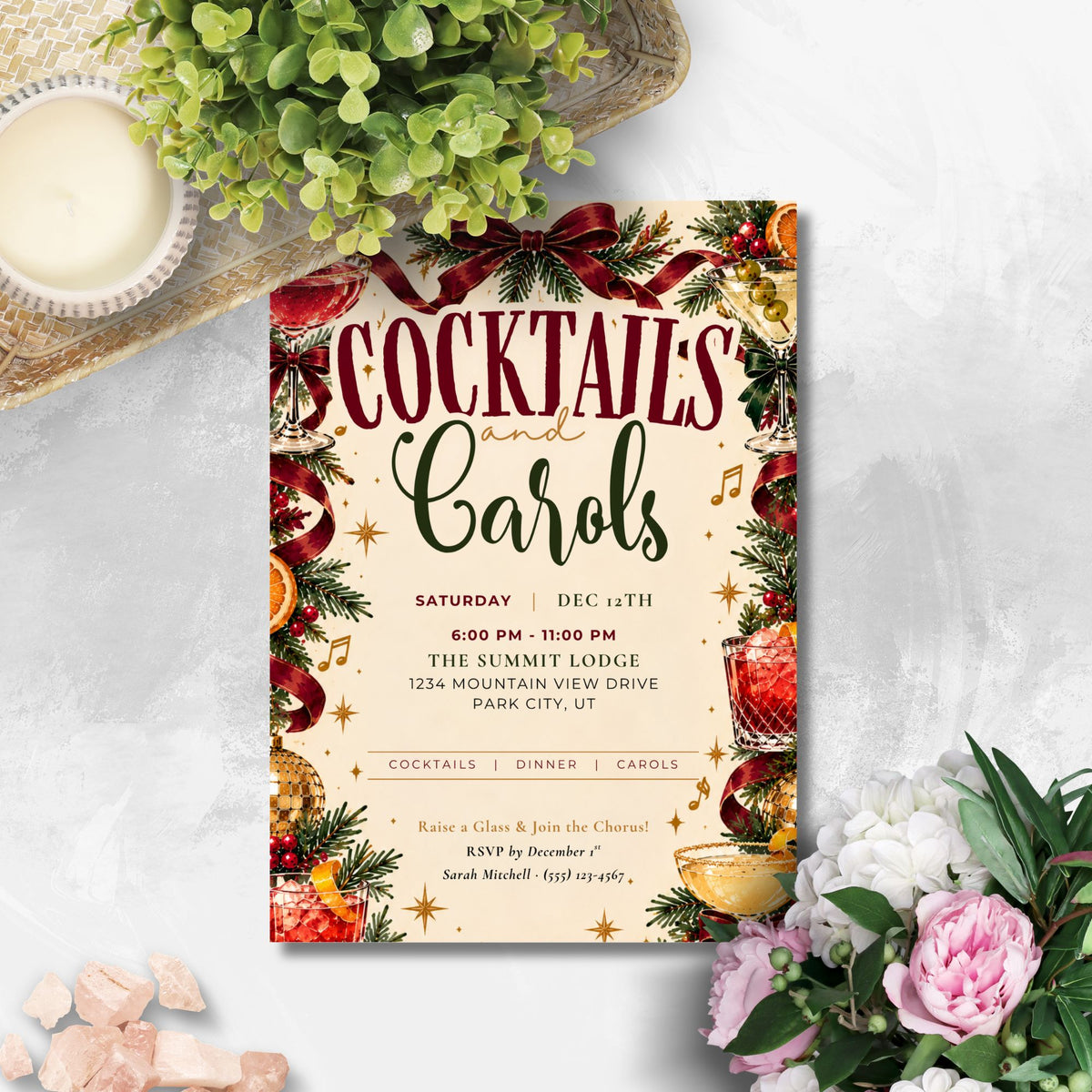 A festive cocktail and carol event invitation card styled in a flat-lay arrangement with green foliage, pink flowers, and a small candle on a light gray textured background.