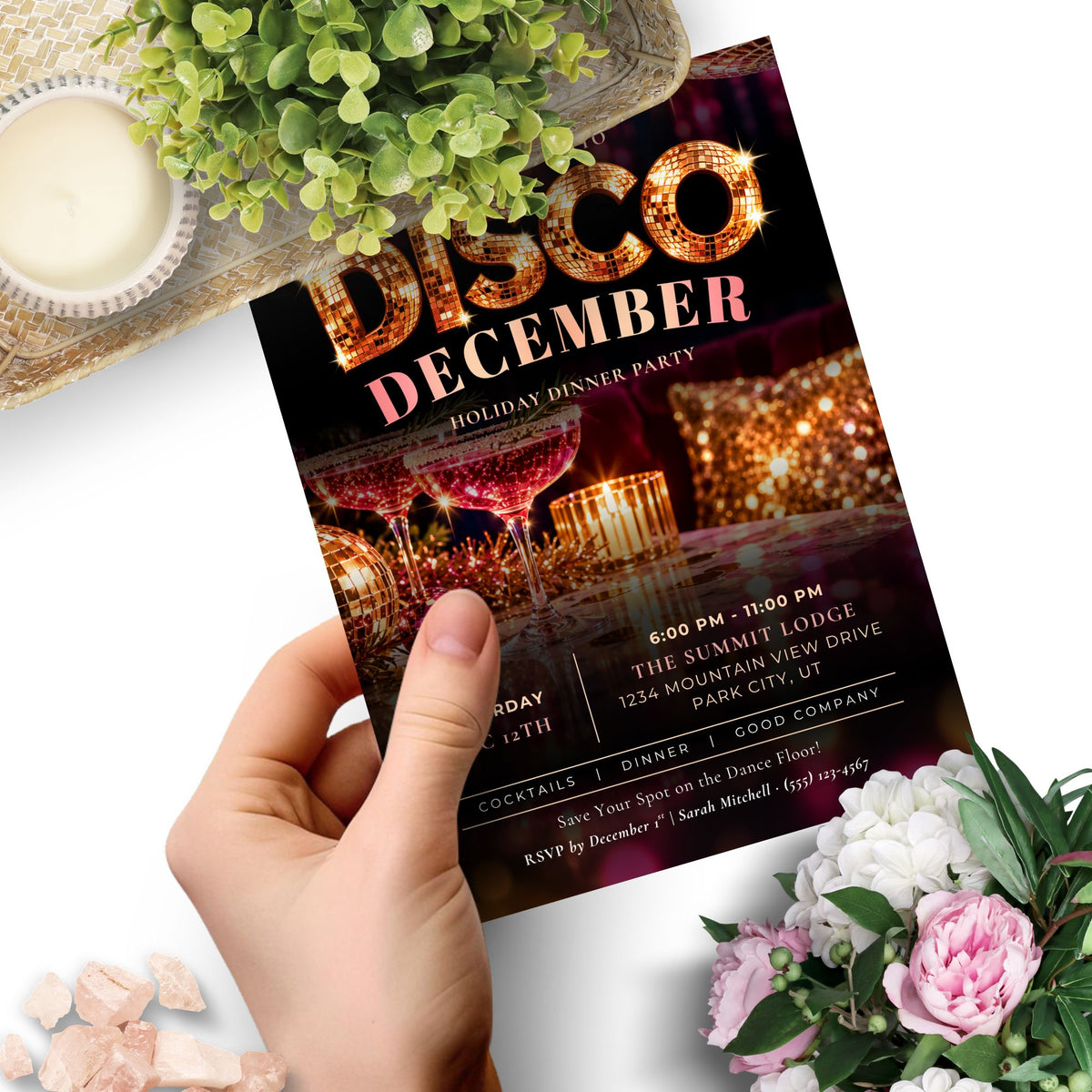 A hand holds a printed invitation card for a 'Disco December' holiday dinner party. The card features a dark background with glittering gold and pink text, and an image of cocktail glasses with red wine and candles.