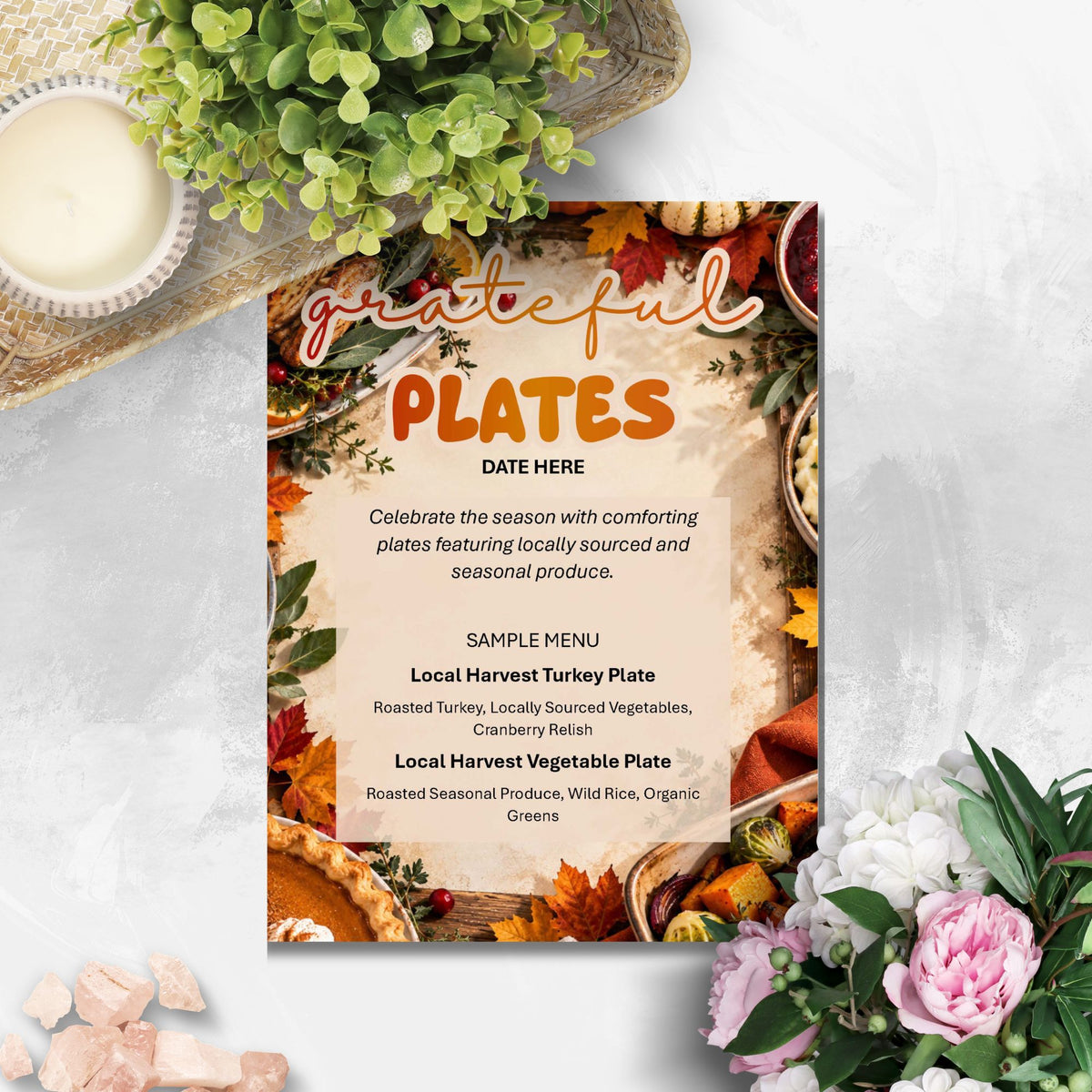 A square printed sign or menu card with autumn-themed food photography around the border and large text reading 'grateful plates' and 'DATE HERE,' styled on a light stone surface with flowers, a candle, and greenery.