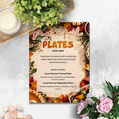 A square printed sign or menu card with autumn-themed food photography around the border and large text reading 'grateful plates' and 'DATE HERE,' styled on a light stone surface with flowers, a candle, and greenery.