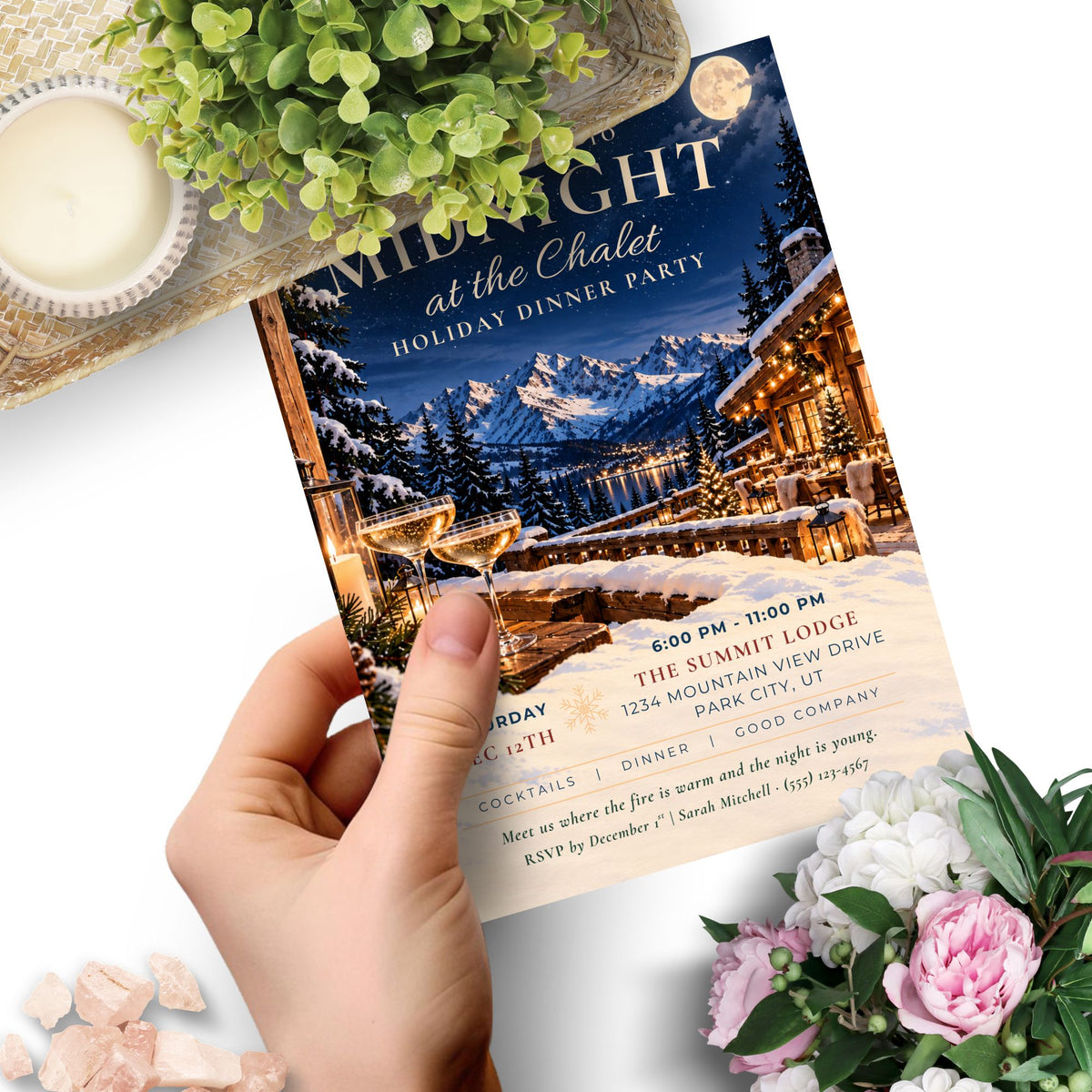 A hand holds a printed invitation card featuring a winter mountain lodge scene with snow, warm lights, and elegant typography. The card reads 'Midnight at the Chalet' and includes event details.