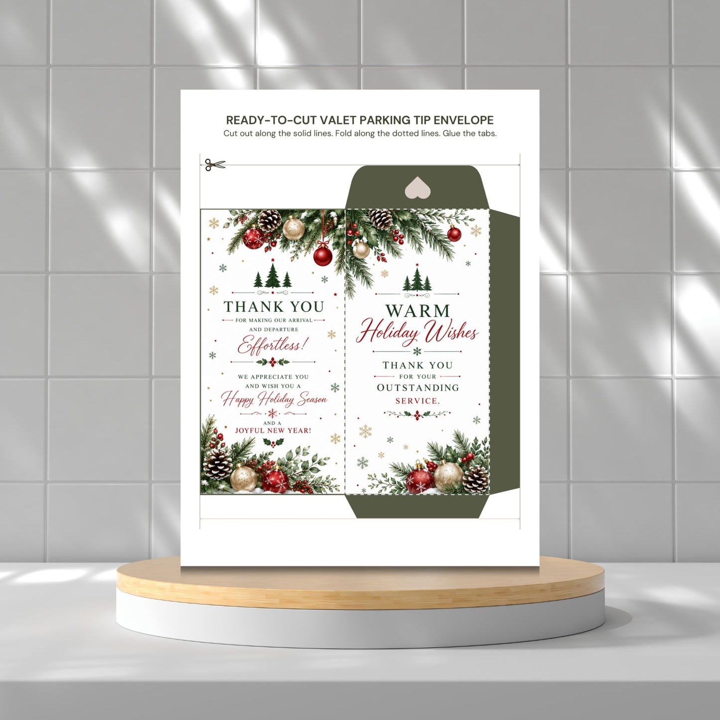 A holiday-themed ready-to-cut valet parking tip envelope displayed on a light wood and white round pedestal against a gray tiled wall. The white envelope features festive green pine garlands with red ornaments, gold pinecones, and snowflake details, along with customizable text reading 'THANK YOU' and 'WARM Holiday Wishes'.