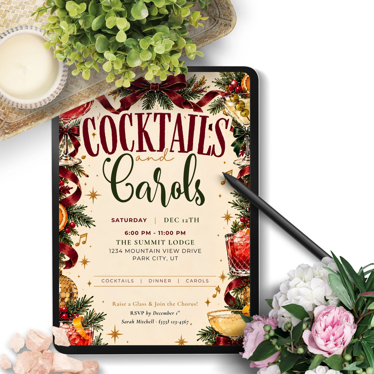 A digital invitation template displayed on a tablet screen, decorated with a festive Christmas theme including pine branches, ornaments, and a cocktail glass. The tablet is styled in a flat-lay setting with a black stylus, a green potted plant, a candle, and pink and white flowers on a white background.