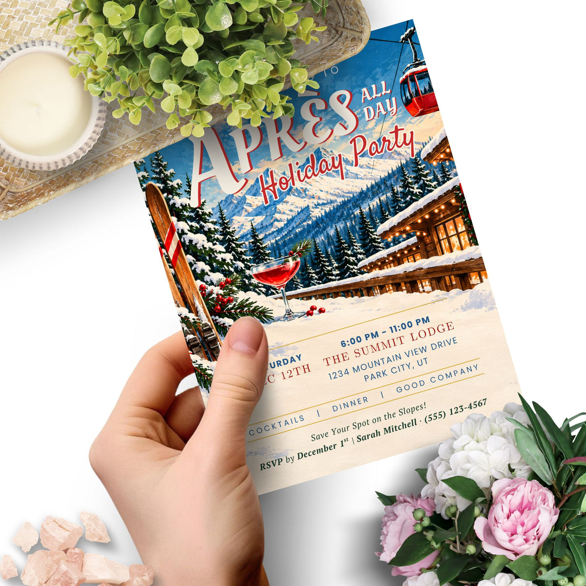 A hand holds a printed invitation card featuring a winter mountain lodge scene with red ski gondolas and the text 'APRES ALL DAY Holiday Party.' The card is styled on a white surface with greenery and flowers around it.