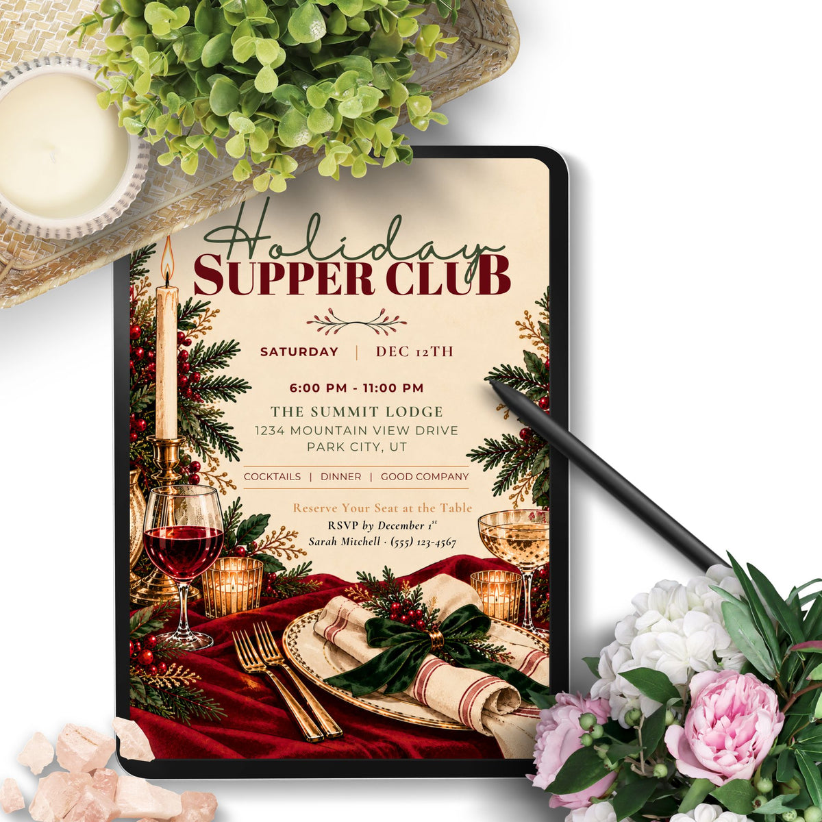 A digital mockup of a holiday supper club invitation displayed on a tablet screen, surrounded by decorative lifestyle props including a candle, greenery, a black stylus, and pink and white flowers on a white background.