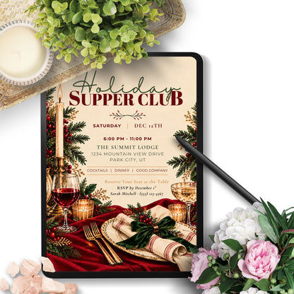 A digital mockup of a holiday supper club invitation displayed on a tablet screen, surrounded by decorative lifestyle props including a candle, greenery, a black stylus, and pink and white flowers on a white background.