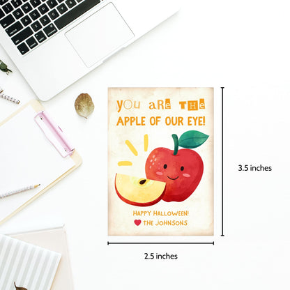 Halloween-themed printable gift tags with the message "You Are the Apple of Our Eye!" featuring a cute apple character perfect for caramel apple gifts. Tags are 2.5 x 3.5 inches, laid out 8 per sheet on a standard 8.5 x 11-inch page. Includes a printable PDF and a PDF with a link to an editable Canva template.