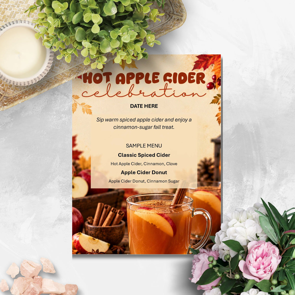 A printed invitation card for a hot apple cider celebration, styled in a flat-lay arrangement with a mug of cider, cinnamon sticks, flowers, and greenery on a light marble surface.