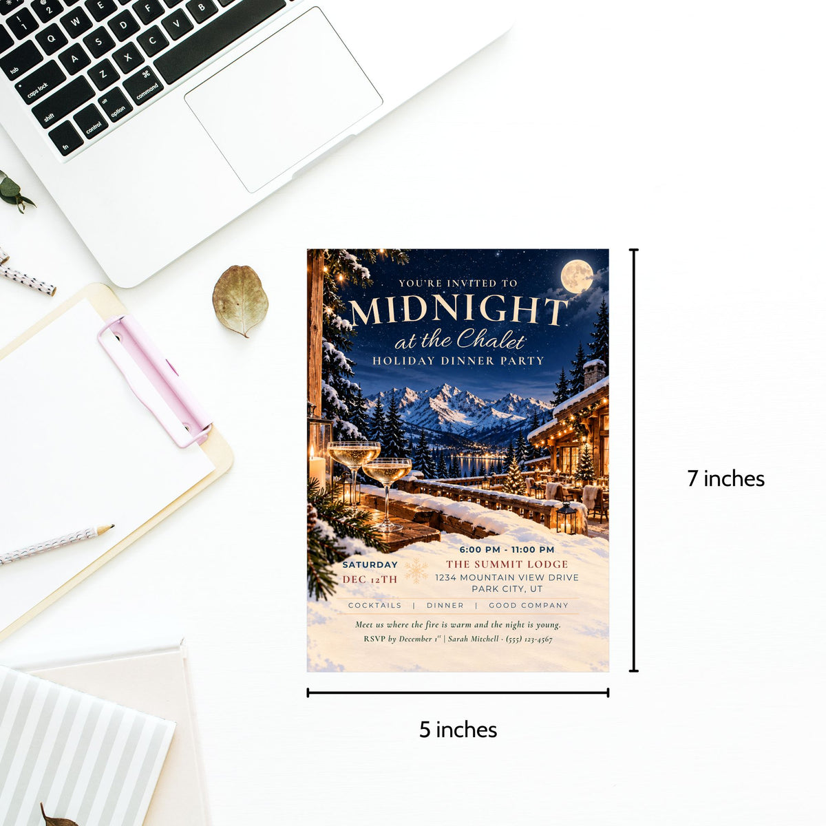 A printed invitation card for a winter holiday dinner party is shown on a white desk with a laptop, notebook, and pen. The card features a scenic mountain cabin illustration at night with snow, warm lights, and a full moon.