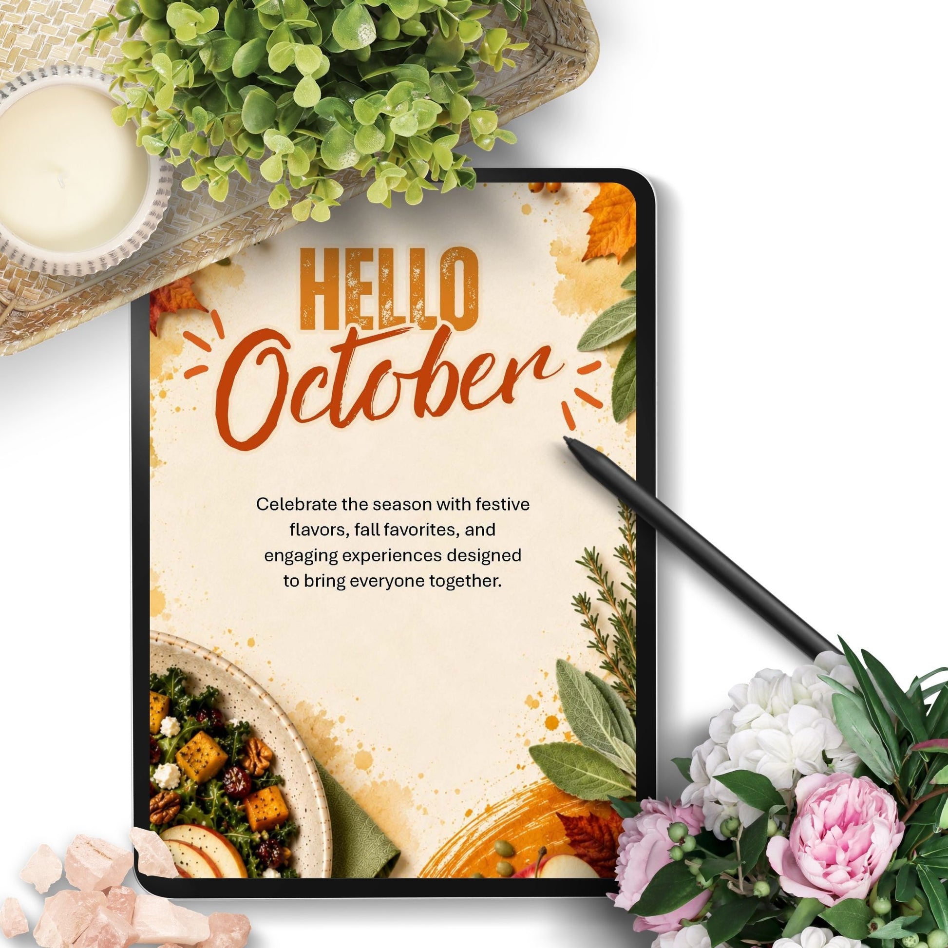 A tablet displaying a 'Hello October' seasonal graphic with food imagery and text, styled on a white background with a black stylus, flowers, greenery, and a small dish.