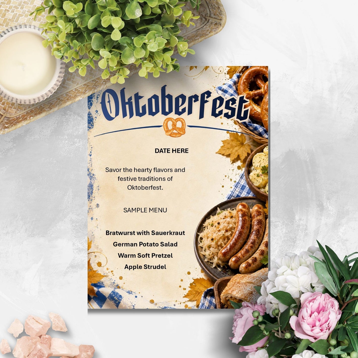 An Oktoberfest-themed printed card or menu cover is shown on a light textured surface, surrounded by decorative flowers, greenery, and a small candle. The card features the word 'Oktoberfest' with a pretzel icon and a sample menu listing beer, sausages, potato salad, pretzel, and apple strudel with food photography along the bottom.