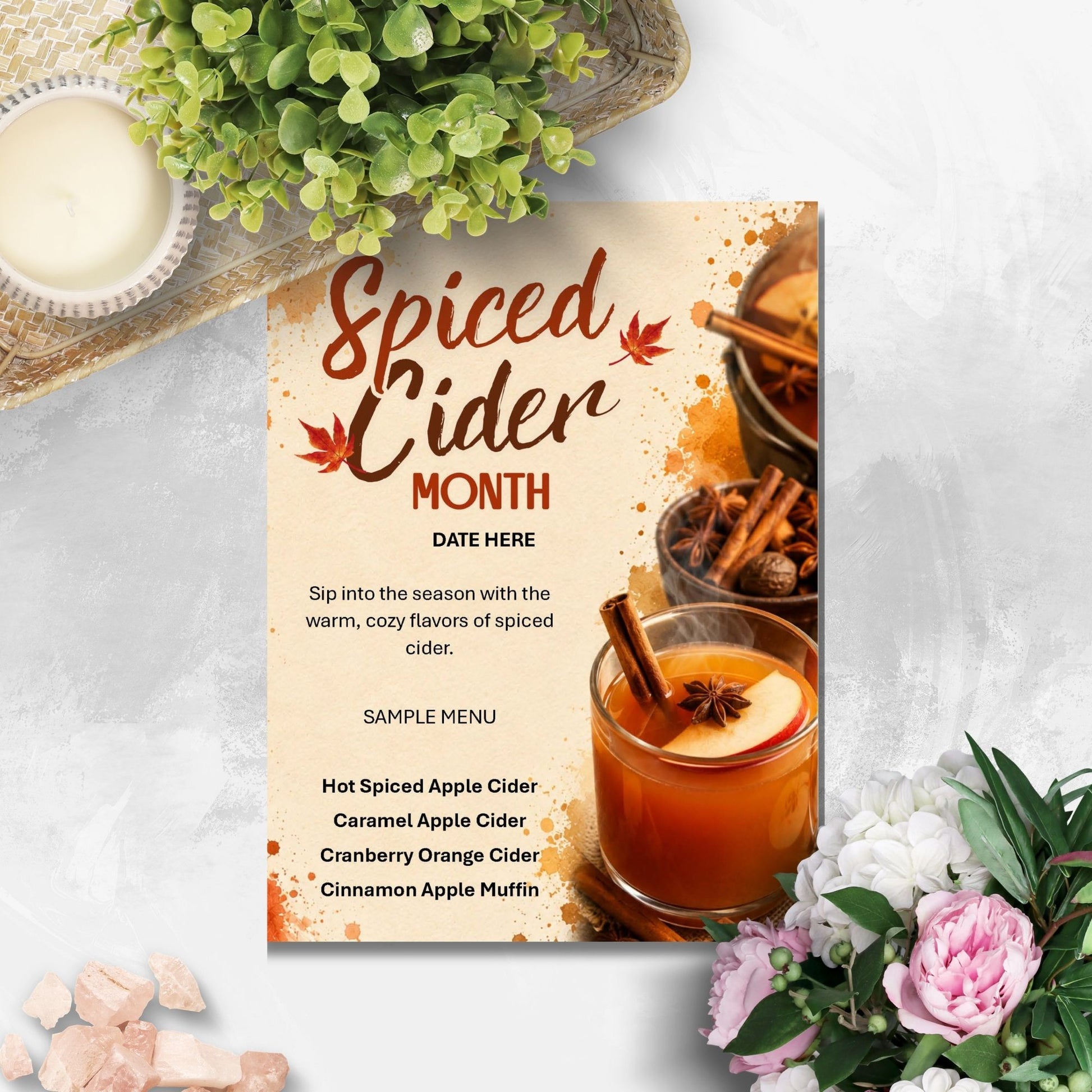 A styled flat-lay featuring a vertical paper sign that reads 'Spiced Cider Month' with a sample menu and cider imagery, surrounded by a candle, greenery, crystals, and pink flowers on a light textured surface.